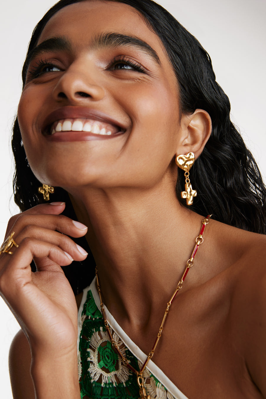 Model shot wearing the soru jewellery textured heart and cross earrings and a red charm chain necklace while leaning forward