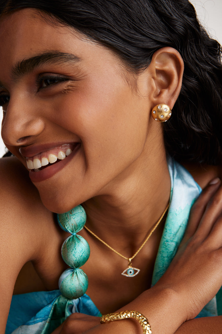 close up model shot side view of round gold star etched earrings with clear crystal embellishment while model is smiling. Model also wearing the mini evil eye charm attached to a rope chain necklace 