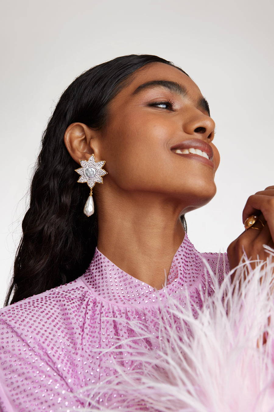 side view of model image shot of large clear crystal embellished flower shaped earrings featuring a round baroque pearl centre and large hanging baroque pearls while model smiling. 