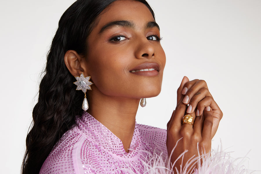 close up model shot of large clear crystal embellished flower shaped earrings featuring a round baroque pearl centre and large hanging baroque pearls. Model also wearing clear crystal embellished round ring while model smiling