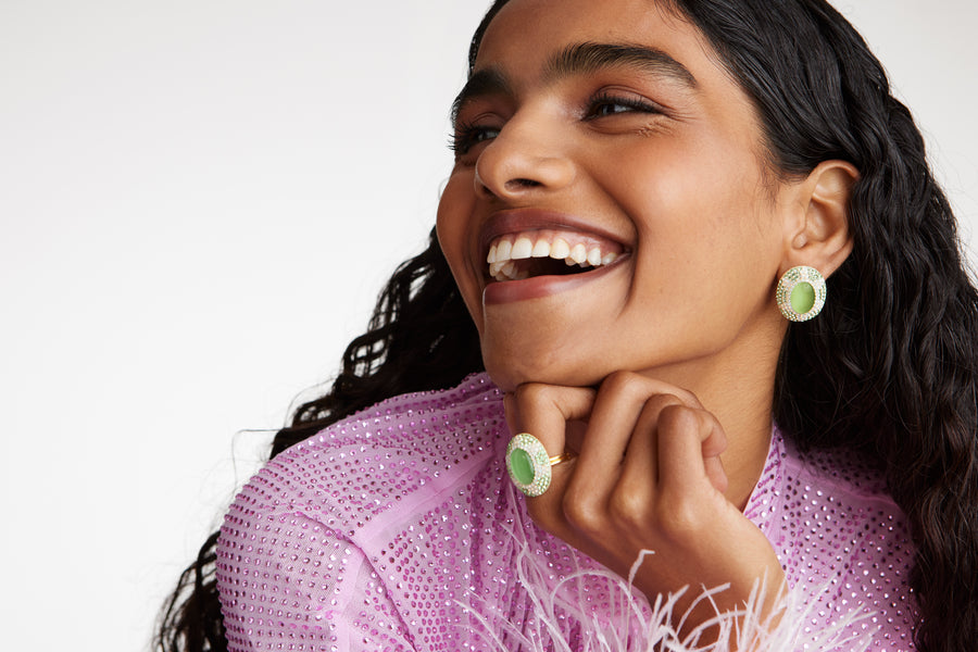 Model shot  wearing soru jewellery oval stud earrings featuring a vibrant green cats eye stone surrounded by sparkling green and opal crystals set with a sun motif. Model also wearing matching ring